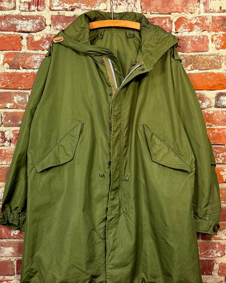 May include: An olive green military-style parka featuring a large hood, a front zipper closure concealed by a snap-button placket, and two large flap pockets on the front. The sleeves have adjustable cuffs with buttons and the hood has a drawstring.