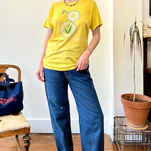 May include: Yellow t-shirt with a daisy graphic and the text "Cookie Champ". The shirt is paired with blue jeans. A navy blue tote bag with "New York" text is on a wooden chair.