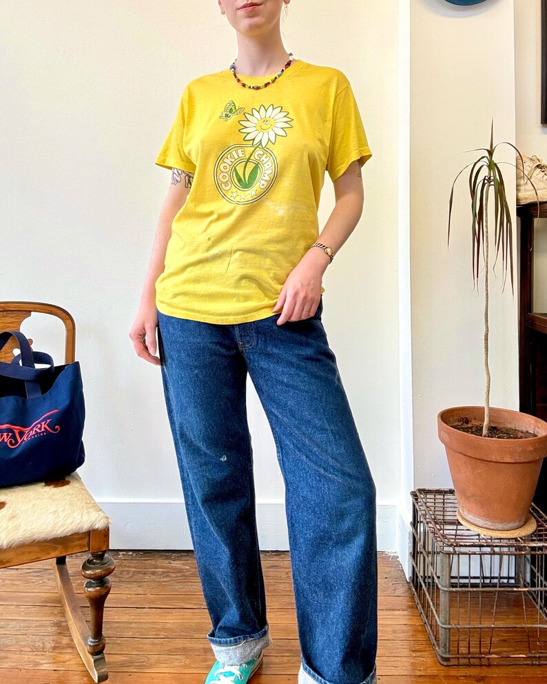 May include: Yellow t-shirt with a daisy graphic and the text "Cookie Champ". The shirt is paired with blue jeans. A navy blue tote bag with "New York" text is on a wooden chair.