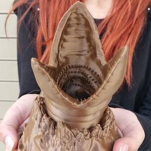 以下が含まれることがあります： 砂虫の口の茶色の3Dプリント彫刻。詳細な歯とテクスチャ加工された表面が特徴です。彫刻は手で持たれており、複雑なデザインと職人技を示しています。ベースは岩のような質感です。