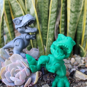May include: Two articulated dinosaur toys, one gray and one green, are positioned among succulents and small rocks. The dinosaurs have open mouths, revealing teeth. The background features tall, green and yellow striped plants.