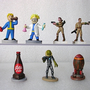 May include: A collection of Fallout-themed figurines. The figures include characters in blue and yellow jumpsuits, a scientist, soldiers, an alien, a Nuka Cola bottle, and a miniature rocket. The figurines are on gray circular bases.