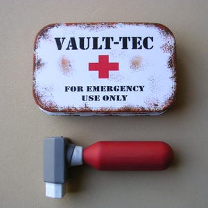 May include: A white metal tin with the words "VAULT-TEC" and "FOR EMERGENCY USE ONLY" printed on it. A red cross is in the center of the tin. A gray and red medical device is next to the tin.