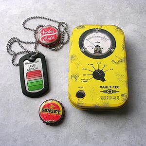 May include: A yellow Vault-Tec radiation meter with a gauge and dial, alongside a black dog tag that reads "RADIATION LEVEL CRITICAL". Also included are two bottle caps, one reading "Nuka Cola" and the other "Sunset Sarsaparilla".