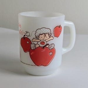 May include: A white milk glass mug featuring a graphic of a character with curly hair sitting on a large red apple, surrounded by smaller red apples. The text Apple Dumplin' is printed in red, along with copyright text at the base of the design.