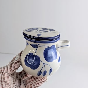 Puede incluir: Un pequeño tarro de cerámica de color crema con tapa a juego, decorado con motivos de frutas y hojas pintados a mano en azul. El tarro tiene un asa corta que sobresale por el lateral y un acabado liso y brillante con bordes azules.