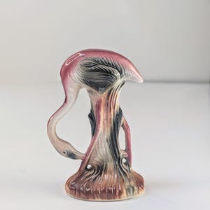 Pode incluir: Uma estatueta de cerâmica de um flamingo com pescoço curvo, apresentando um acabamento brilhante em tons de rosa, castanho e preto. A ave está representada sobre uma base texturizada semelhante a uma árvore com detalhes de pinceladas.