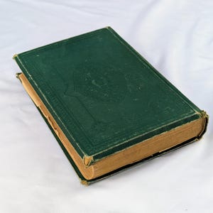 May include: A vintage hardcover book with a dark green cloth cover featuring an embossed decorative border and central emblem. The book has aged, tan-colored pages visible along the edges, with slight wear on the corners and spine edges.
