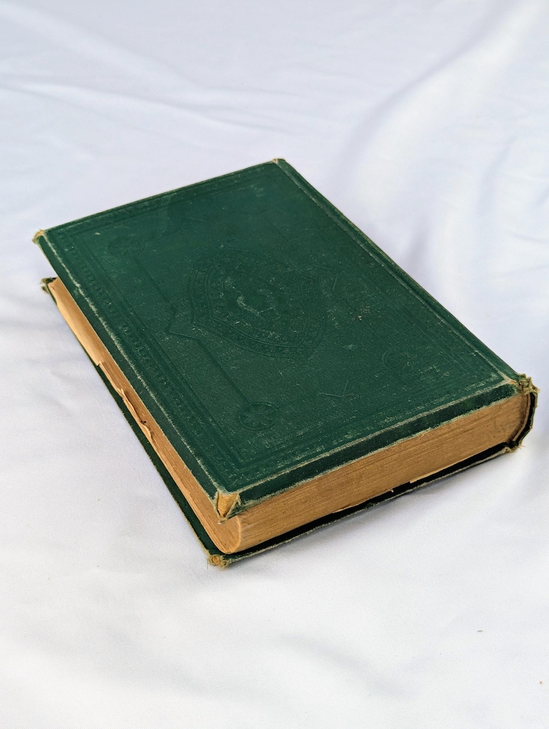May include: A vintage hardcover book with a dark green cloth cover featuring an embossed decorative border and central emblem. The book has aged, tan-colored pages visible along the edges, with slight wear on the corners and spine edges.
