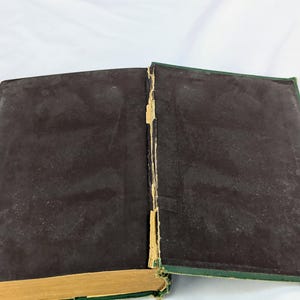 May include: An open hardcover book with dark brown endpapers and a green exterior cover. The central spine area shows visible wear with frayed, light-colored binding material exposed between the two covers and the spine edge of the book.