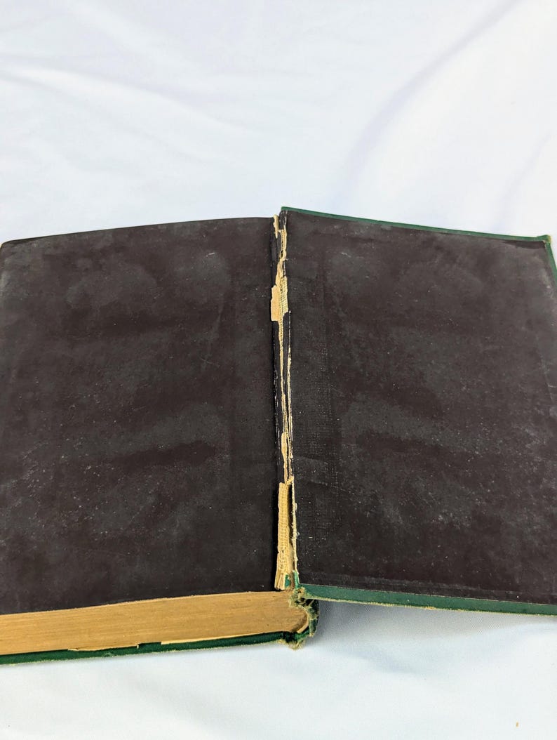 May include: An open hardcover book with dark brown endpapers and a green exterior cover. The central spine area shows visible wear with frayed, light-colored binding material exposed between the two covers and the spine edge of the book.