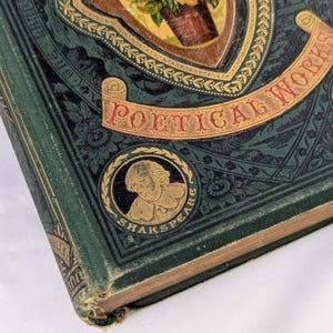 May include: A dark green hardcover book titled Poetical Works with an ornate gold-embossed floral illustration on the cover. A circular medallion features a portrait of William Shakespeare with the text SHAKSPEARE inscribed around the border.