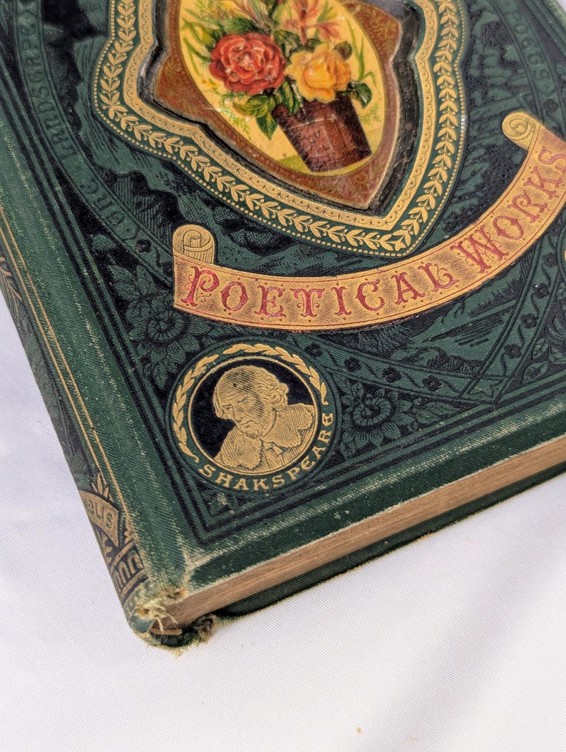 May include: A dark green hardcover book titled Poetical Works with an ornate gold-embossed floral illustration on the cover. A circular medallion features a portrait of William Shakespeare with the text SHAKSPEARE inscribed around the border.