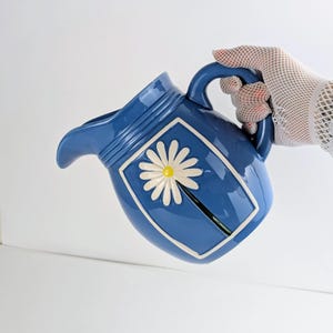 May include: A glossy blue ceramic pitcher featuring a raised white daisy flower with a yellow center. The pitcher has a curved handle, a wide pouring spout, and a rectangular embossed frame surrounding the floral design on its side.