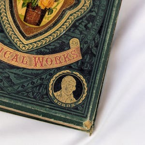 May include: A dark green hardcover book titled POETICAL WORKS with gold embossed lettering and decorative borders. The cover features a central oval illustration of a flower bouquet and a circular portrait of a man labeled BURNS at the bottom.
