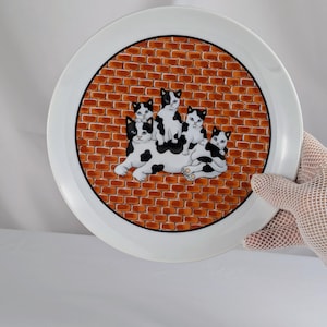 May include: A round white ceramic plate featuring a central circular illustration of five black and white cats huddled together against a background of orange-brown brickwork. The cats are depicted in various poses, with one large cat lying down.