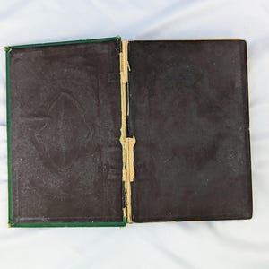 May include: An open, vintage hardcover book with dark brown, textured endpapers featuring a subtle embossed pattern. The spine area shows visible wear, with frayed tan fabric and exposed binding material between the two dark covers.