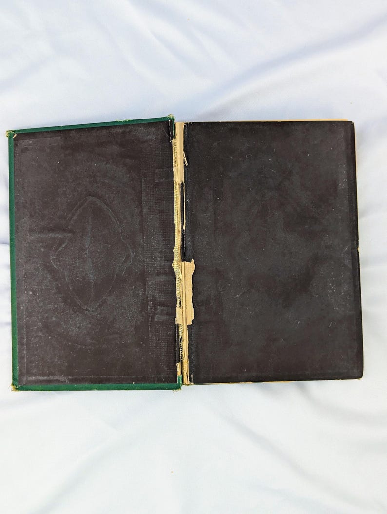 May include: An open, vintage hardcover book with dark brown, textured endpapers featuring a subtle embossed pattern. The spine area shows visible wear, with frayed tan fabric and exposed binding material between the two dark covers.