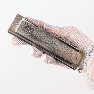 May include: A silver-toned M. Hohner Super Chromonica chromatic harmonica featuring an ornate floral engraving on the top cover plate. The metal instrument displays the text Super Chromonica, Chromatic, and M. Hohner, with numbered holes 1 through 12.