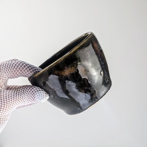 May include: A small, hand-thrown ceramic cup with a glossy, dark black glaze. The surface features subtle, mottled brown and tan variations near the rim and base, creating a reflective, uneven texture on the exterior of the rounded vessel.