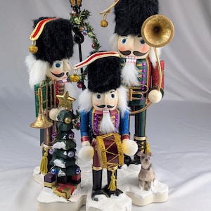 May include: Three nutcracker figures in military-style uniforms with black fuzzy hats, white beards, and gold accents. One plays a trumpet, one a horn, and one a drum. They stand on a snowy base with a small Christmas tree, gifts, and a small dog.