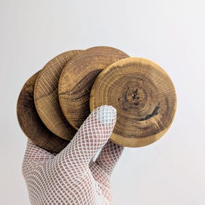 May include: A set of four circular wooden coasters featuring visible natural grain patterns and dark brown tree ring details. Each coaster is crafted from a slice of wood with a smooth finish, showcasing the organic texture and light brown tones.