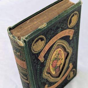 May include: A vintage hardcover book with a dark green cloth cover featuring gold embossed lettering and decorative borders. The front cover displays a central oval illustration of a floral bouquet, flanked by two circular gold-toned architectural vignettes.