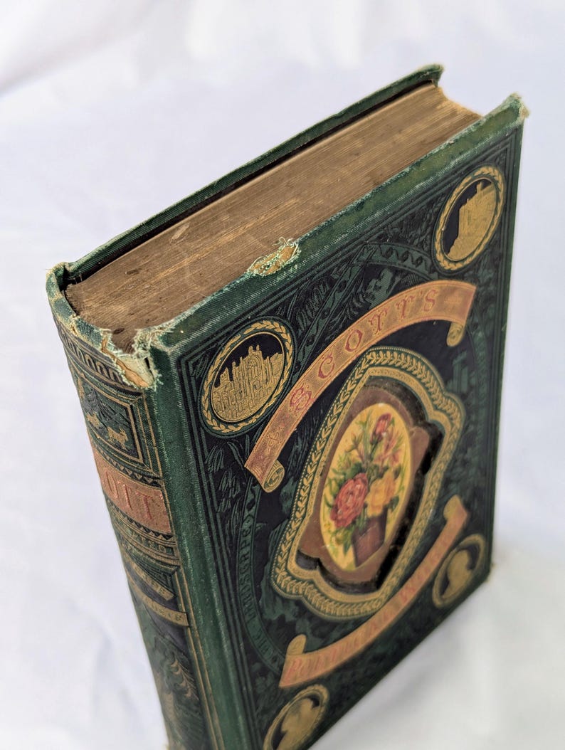 May include: A vintage hardcover book with a dark green cloth cover featuring gold embossed lettering and decorative borders. The front cover displays a central oval illustration of a floral bouquet, flanked by two circular gold-toned architectural vignettes.