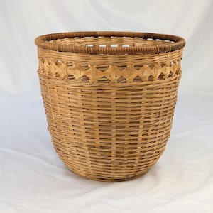 May include: A round, light brown woven basket featuring a wide, open top and a decorative crisscross pattern near the rim. The basket is constructed from thin, flexible strips of natural material, showcasing a consistent, vertical weave design.