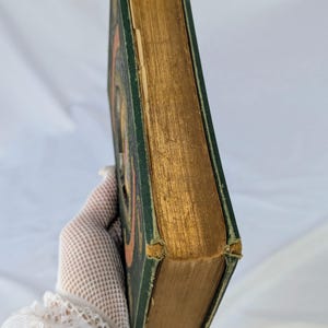 May include: A hardcover book with a dark green cover and gold-gilded page edges. The book features a visible orange circular design on the front cover. The corners of the green cover show signs of wear and fraying, revealing the underlying material.