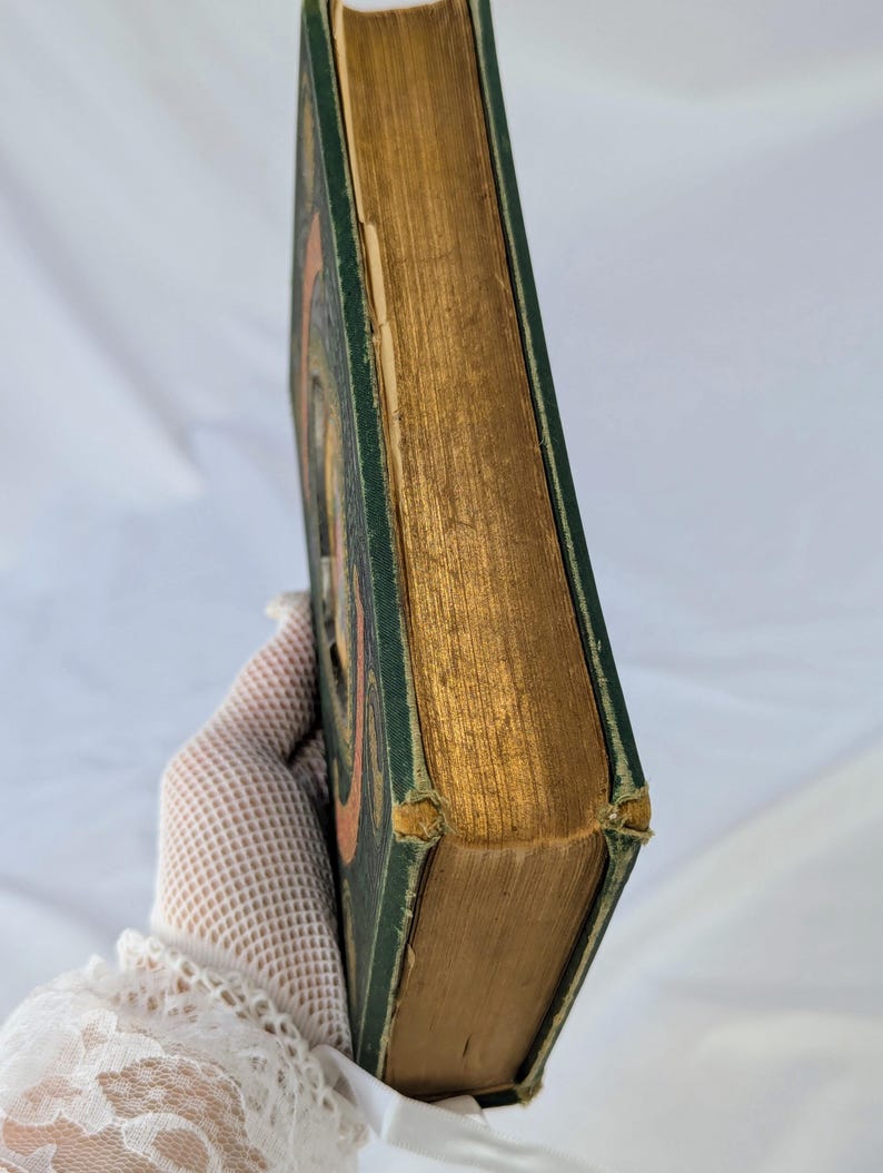 May include: A hardcover book with a dark green cover and gold-gilded page edges. The book features a visible orange circular design on the front cover. The corners of the green cover show signs of wear and fraying, revealing the underlying material.