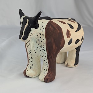 May include: A hand-painted ceramic figurine of a bull with a cream-colored base. The bull features a black head, dark brown patches on the body, and black spots on the chest and rear. It stands on four sturdy legs with a smooth, matte finish.