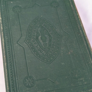 May include: A dark green hardcover book featuring an embossed central emblem of a lyre surrounded by decorative text. The cover includes a geometric border with circular floral motifs at each corner, showing signs of wear on the edges.