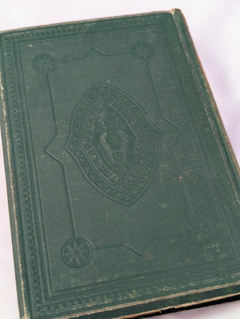 May include: A dark green hardcover book featuring an embossed central emblem of a lyre surrounded by decorative text. The cover includes a geometric border with circular floral motifs at each corner, showing signs of wear on the edges.