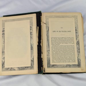 May include: An open antique book titled The Life of Sir Walter Scott with aged, cream-colored pages. The left page features a decorative border, while the right page contains printed text detailing the biography of Sir Walter Scott.