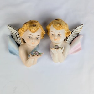 May include: Two ceramic wall plaques shaped like cherubs with blonde hair and white wings. One cherub has a light blue drape and holds a small purple flower, while the other has a light pink drape and also holds a small purple flower in their hands.