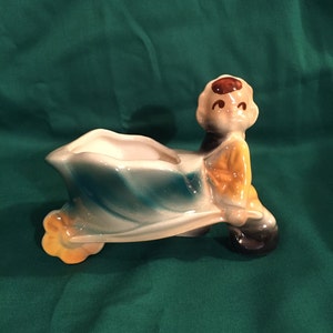 Op de afbeelding: Een keramische plantenbak in de vorm van een persoon die een kruiwagen duwt. De plantenbak is geschilderd in tinten blauw, groen, geel en bruin. De persoon heeft een bruin gezicht met een geschilderde glimlach en bruin haar.