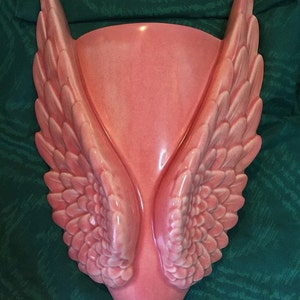 May include: A pink ceramic wall hanging shaped like a pair of angel wings. The wings are detailed with individual feathers and curve upwards. The background is a dark green fabric.