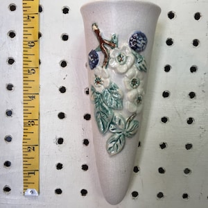 May include: White ceramic wall vase with a floral design. The vase is shaped like a cone and has a hanging loop at the top. The flowers are white with green leaves and blue berries.