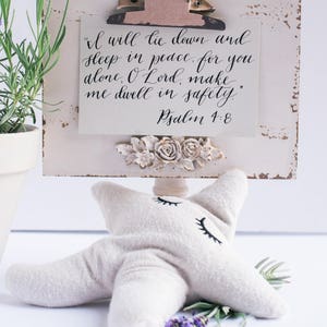 May include: A white wooden sign with a distressed finish, featuring a clip holding a card with the text "I will lie down and sleep in peace, for you alone, O Lord, make me dwell in safety." Psalm 4:8. The sign is decorated with a floral design and is sitting on a white star-shaped pillow.