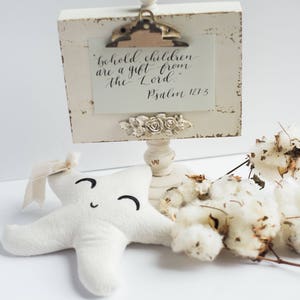 May include: A white wooden sign with a distressed finish, featuring a quote about children being a gift from the Lord, with the verse Psalm 127:3. The sign is on a white pedestal with a floral design. A white plush star with a smiling face is in the foreground, along with dried cotton bolls.