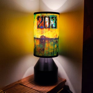 Pode incluir: Um abajur de mesa preto com um abajur cilíndrico que apresenta um design abstrato amarelo, verde e preto com o número "203" impresso nele.