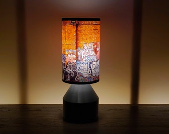 May include: A cylindrical table lamp with a graffiti-style lampshade. The shade features a brick wall background covered in colorful graffiti art. The base is a dark gray color.