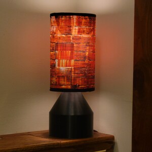 May include: A black table lamp with a cylindrical lampshade featuring a brick pattern in shades of orange and brown. The lamp is turned on and emitting a warm glow.