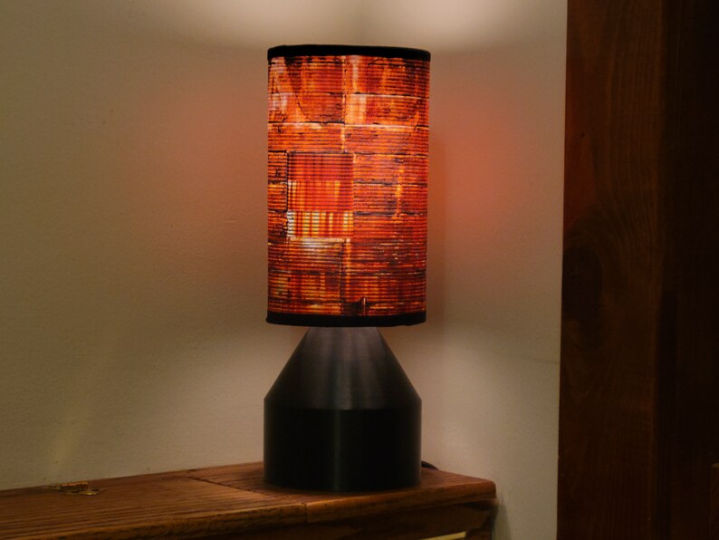 May include: A black table lamp with a cylindrical lampshade featuring a brick pattern in shades of orange and brown. The lamp is turned on and emitting a warm glow.