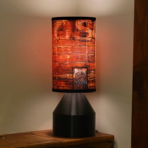 May include: A table lamp with a black base and a cylindrical lampshade. The lampshade has a rusty metal design.