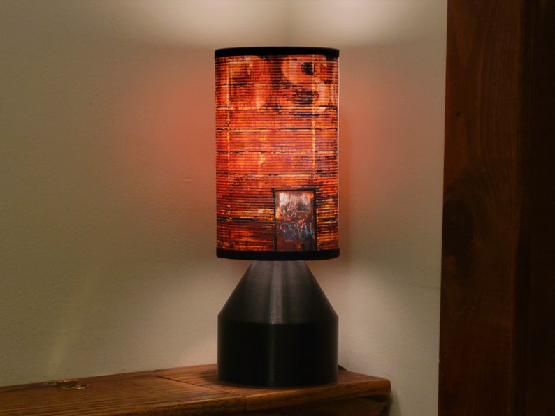 May include: A table lamp with a black base and a cylindrical lampshade. The lampshade has a rusty metal design.