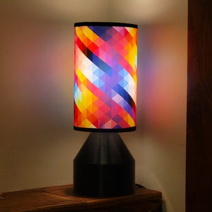 May include: A table lamp with a black base and a colorful geometric patterned lampshade. The lampshade has a mix of red, yellow, blue, and white colors.
