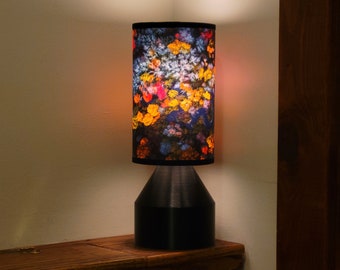 Pode incluir: Um abajur de mesa preto com um abajur estampado com flores. O abajur apresenta uma variedade de cores, incluindo amarelo, laranja, vermelho e azul. A lâmpada está sobre uma superfície de madeira.