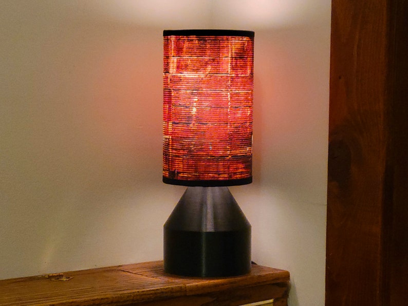 May include: A black table lamp with a red and orange patterned lampshade. The lamp is turned on and the light is shining through the lampshade.